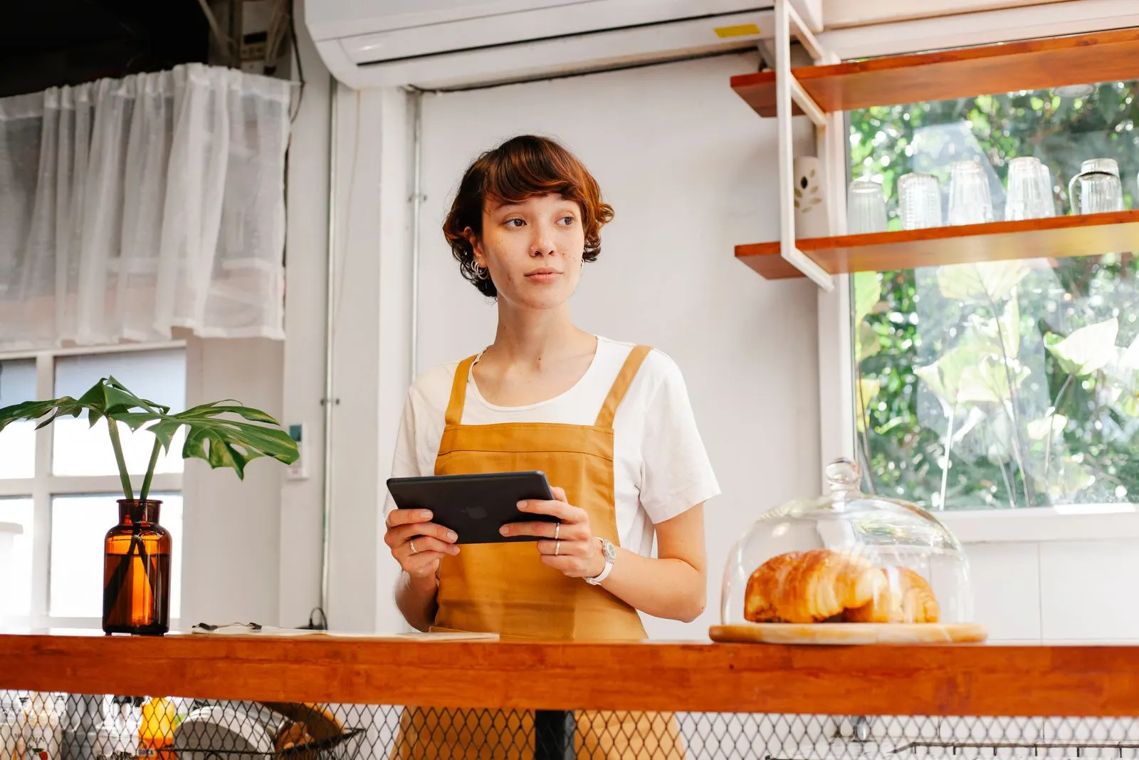 Cafe worker in an orange apron holding a tablet behind a wooden counter with a covered plate of croissants