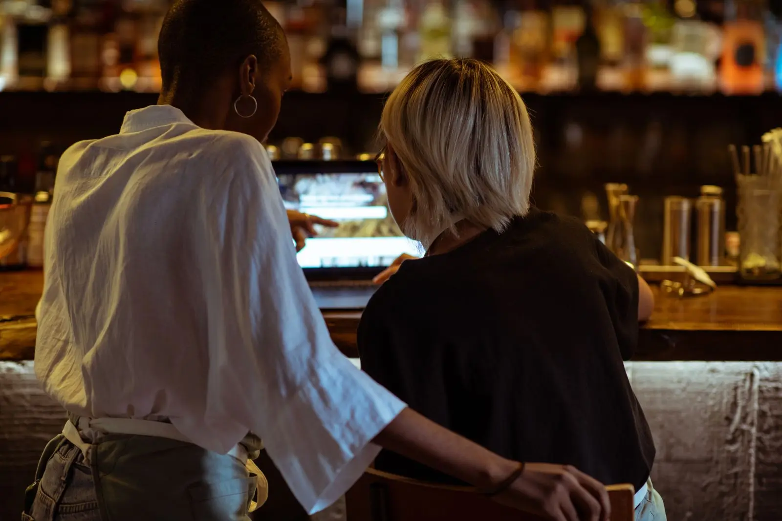 Two restaurant workers at a bar counter looking at a website on a laptop, seen from behind