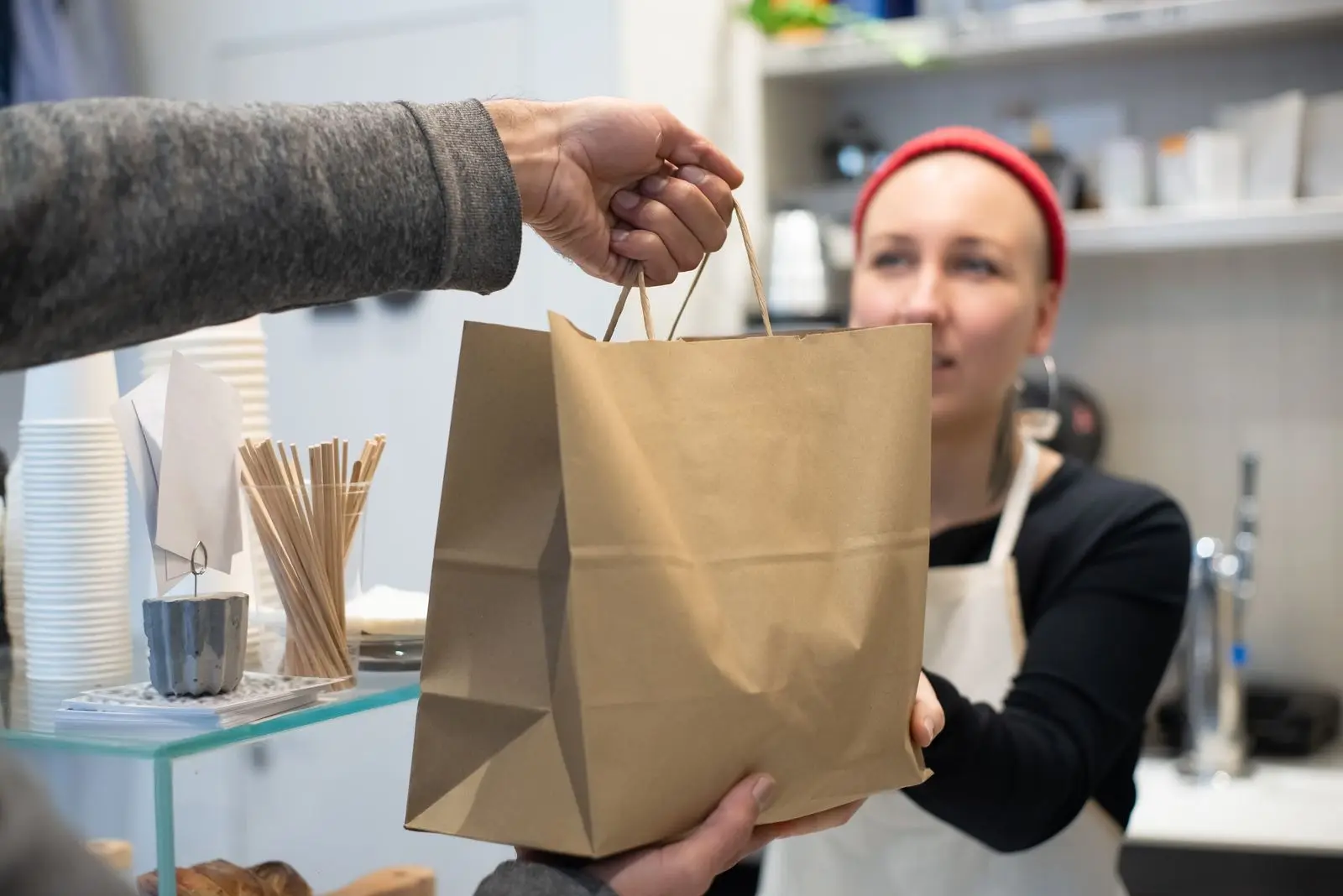 Restaurant worker in an apron handing a brown paper takeout bag across the counter to a customer